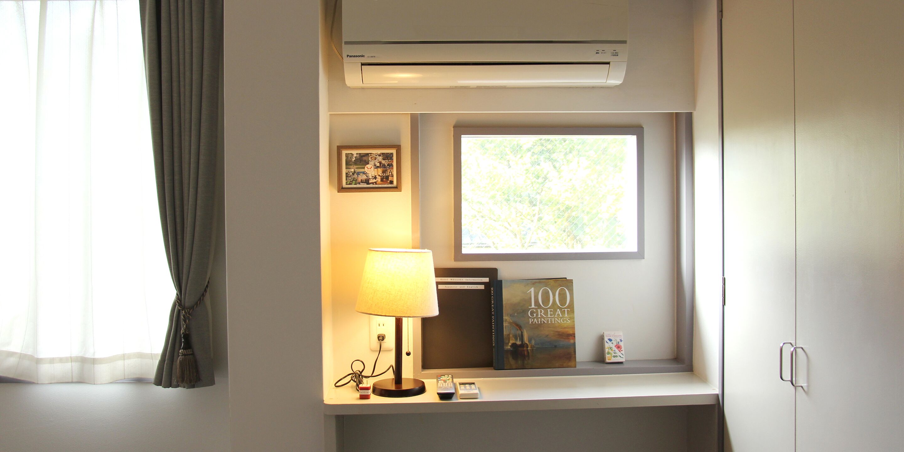 A hotel room with a window, an air conditioner, a lamp, a book titled '100 Great Paintings', and a small decorative item on the windowsill.