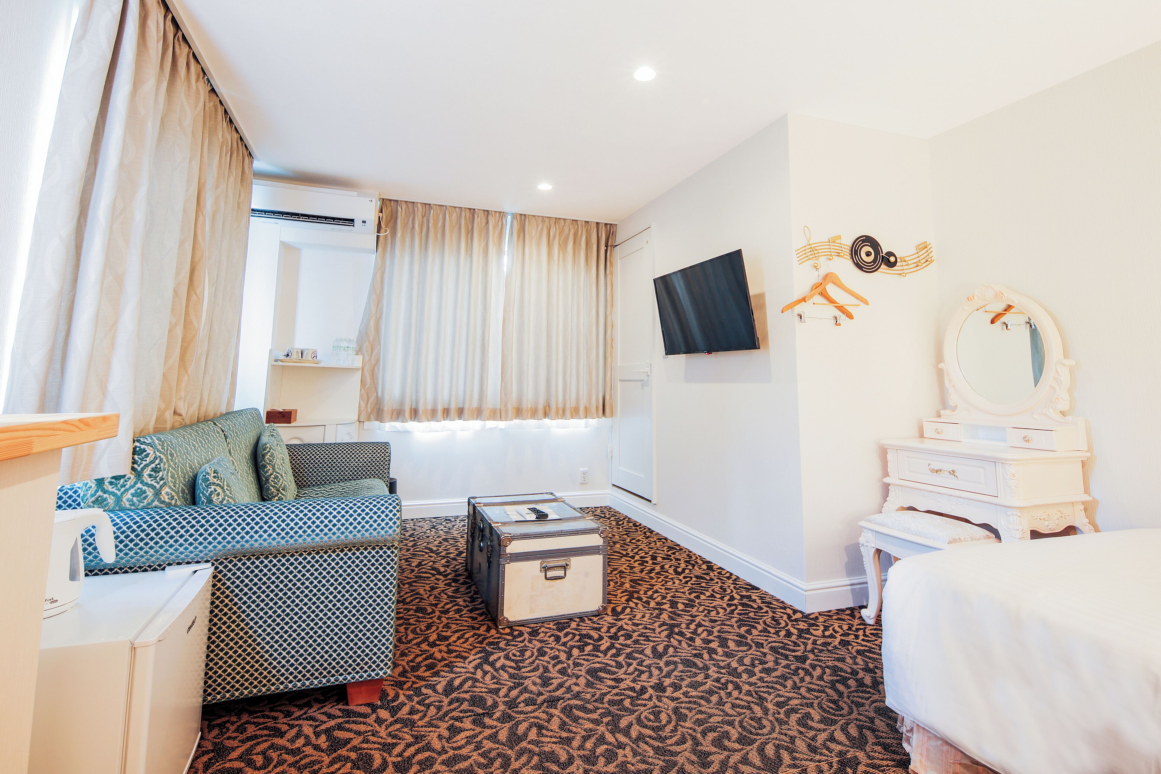 A hotel room with a patterned carpet, a sofa, a small table, a dresser, a mirror, and a television on the wall.