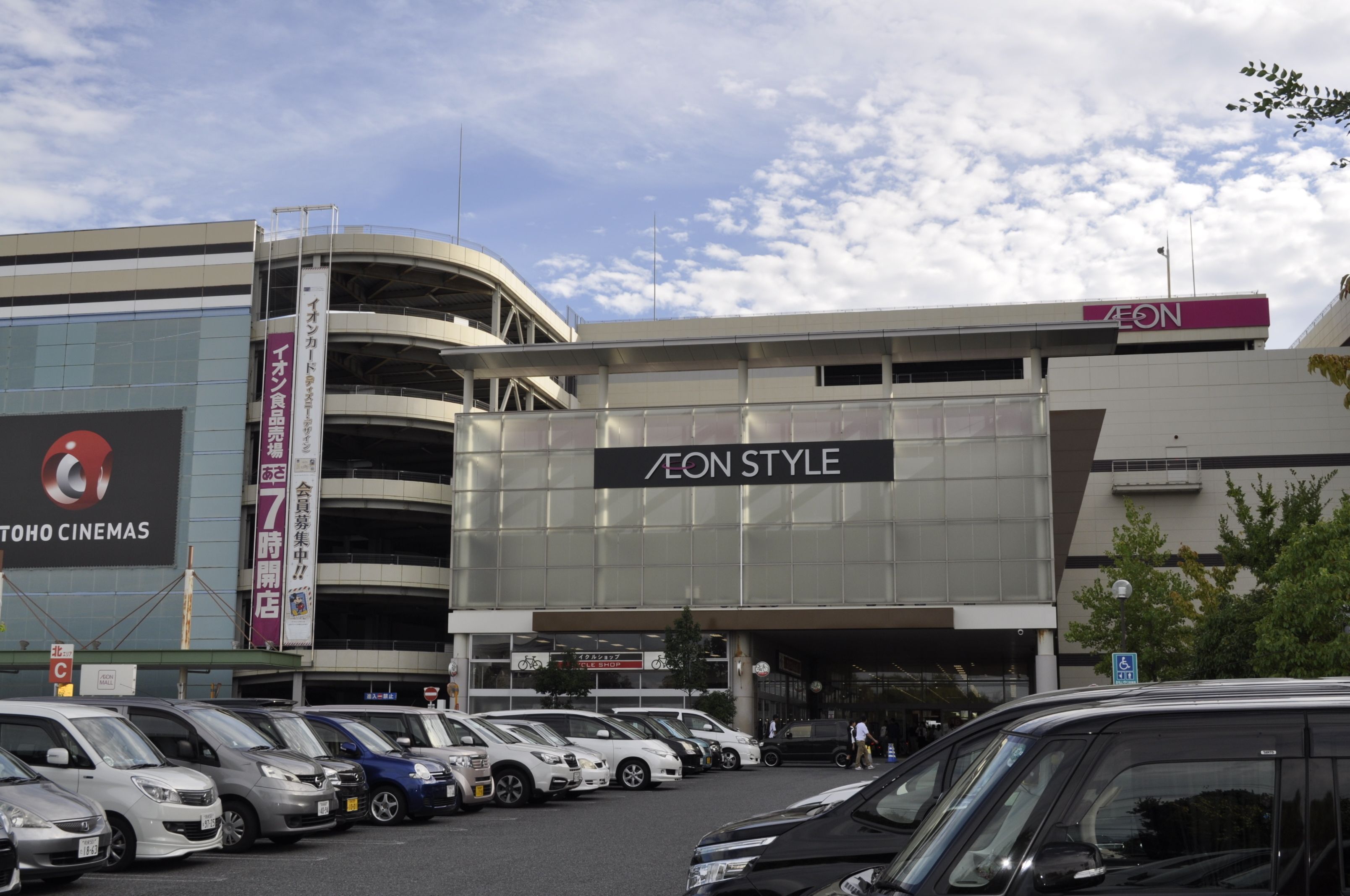 Un estacionamiento con varios automóviles, un edificio con el rótulo 'AEON STYLE' y un cartel de TOHO CINEMAS.