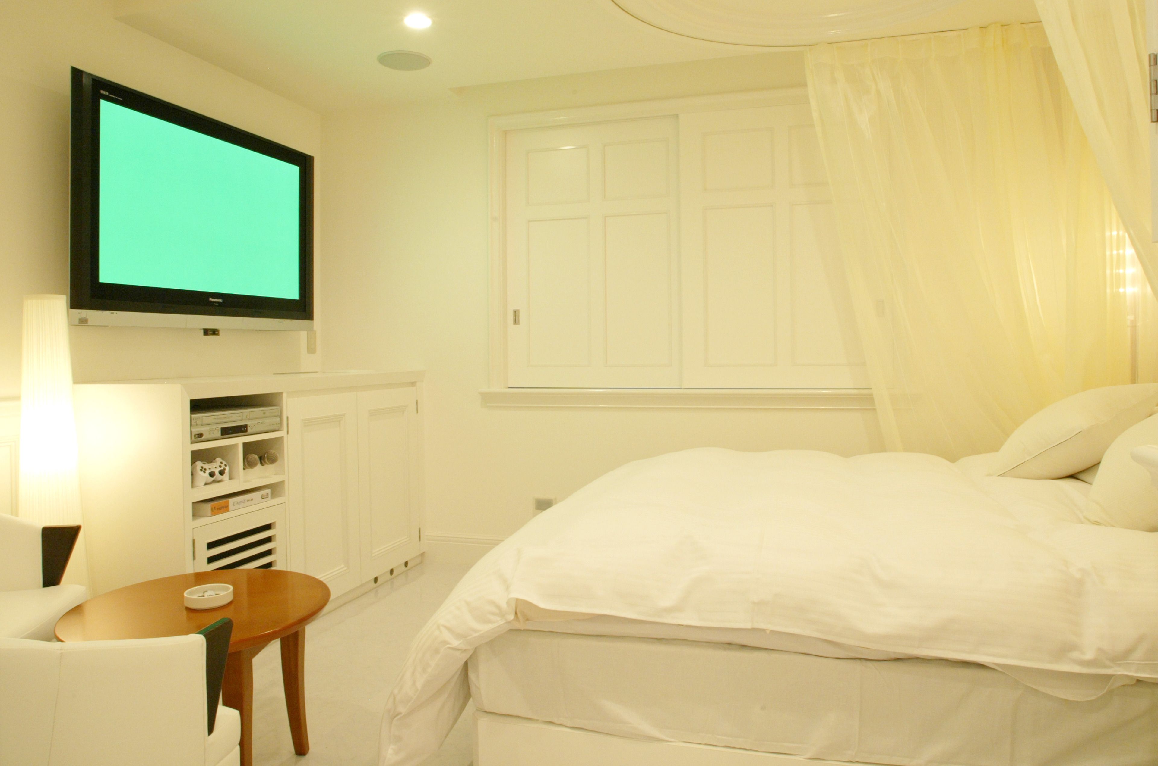 A bedroom with a bed, a television mounted on the wall, a bedside table, and a window with curtains.