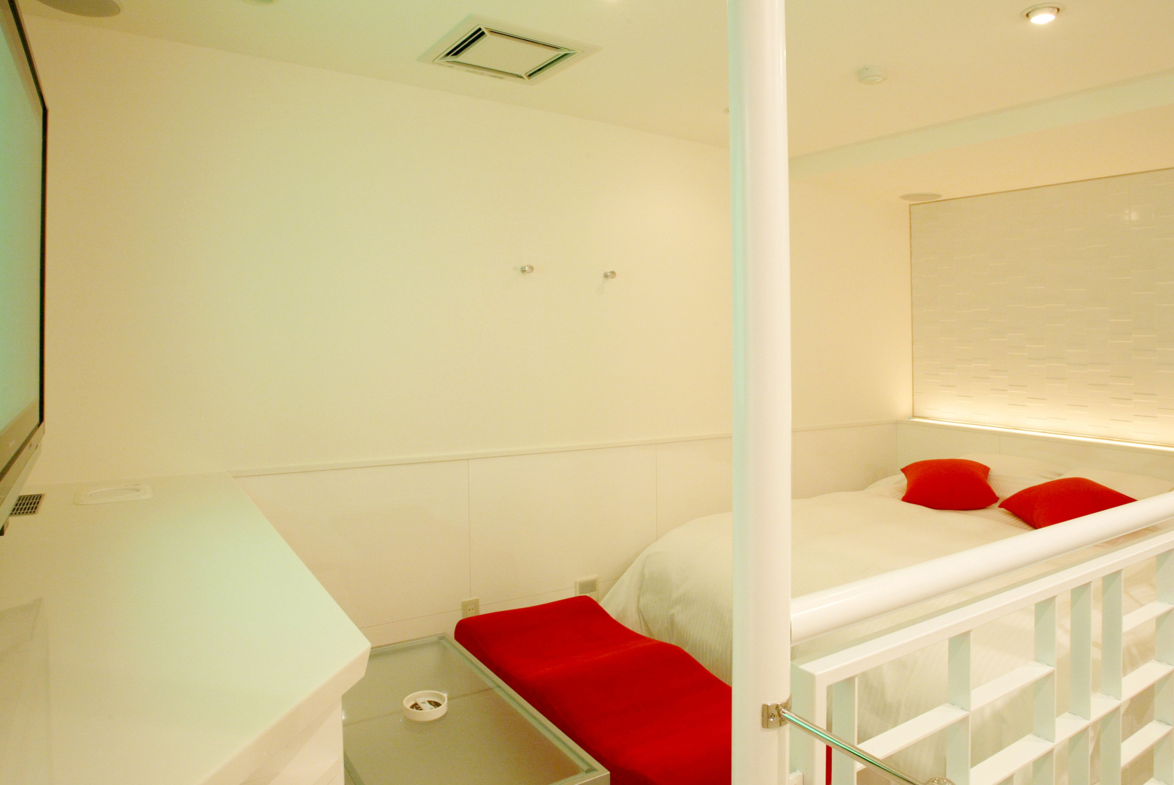 Habitació d'hotel petita i blanca, d'una sola planta, amb un llit, una banca vermella i una barana blanca.