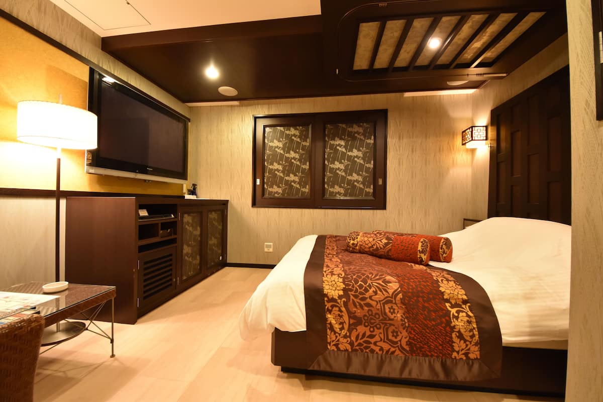 A hotel room with a bed, a TV mounted on the wall, a desk with a lamp, and a window with decorative shutters.