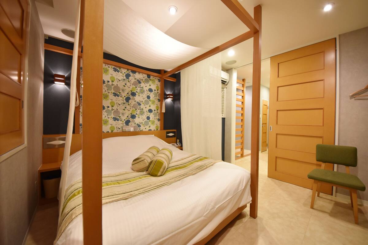 A bedroom with a four-poster bed, a green chair, a wooden wardrobe, and a floral-patterned wall.