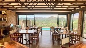 A spacious dining area with multiple wooden tables and chairs, large windows offering a view of mountains, and a brick wall with a fireplace. - Windhoek Game Camp (Windhoek)