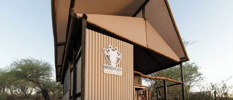 A windhoek game camp structure with a corrugated metal exterior, a wooden deck, and a large canopy.