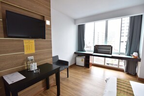 A hotel room with a wooden floor, a desk with a computer, a TV mounted on the wall, a bed with a green and white striped blanket, and a window with curtains. - Mayflower Grande Hotel Hat Yai (Hat Yai)