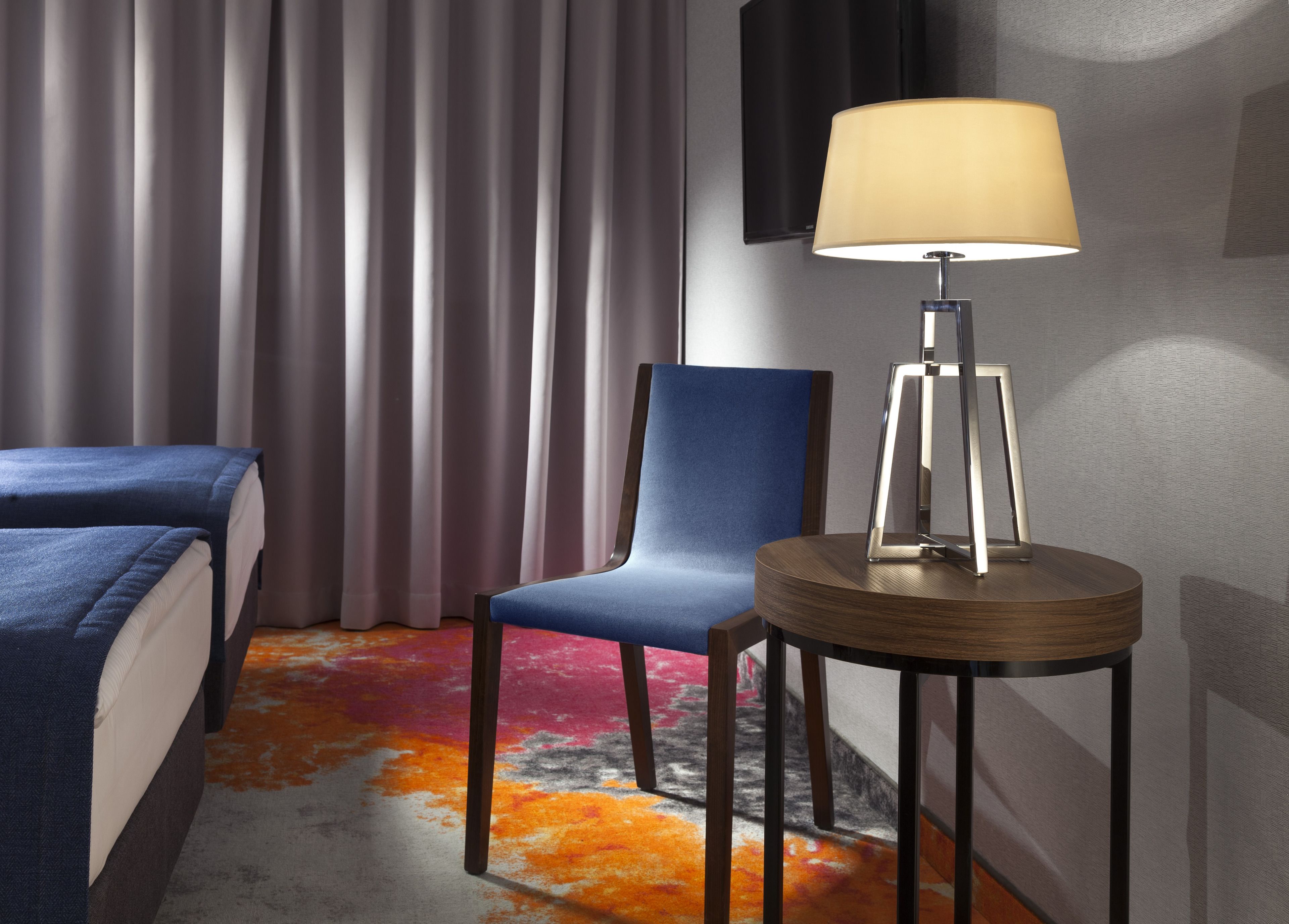 Ett hotellrum med en säng, en blå stol, ett runt bord med en lampa och en färgglad matta.