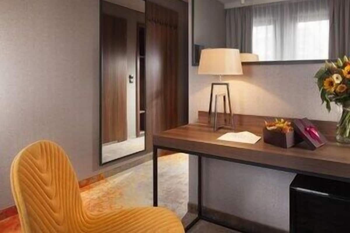 A hotel room with a wooden desk, a chair, a lamp, a vase with flowers, and a mirror.