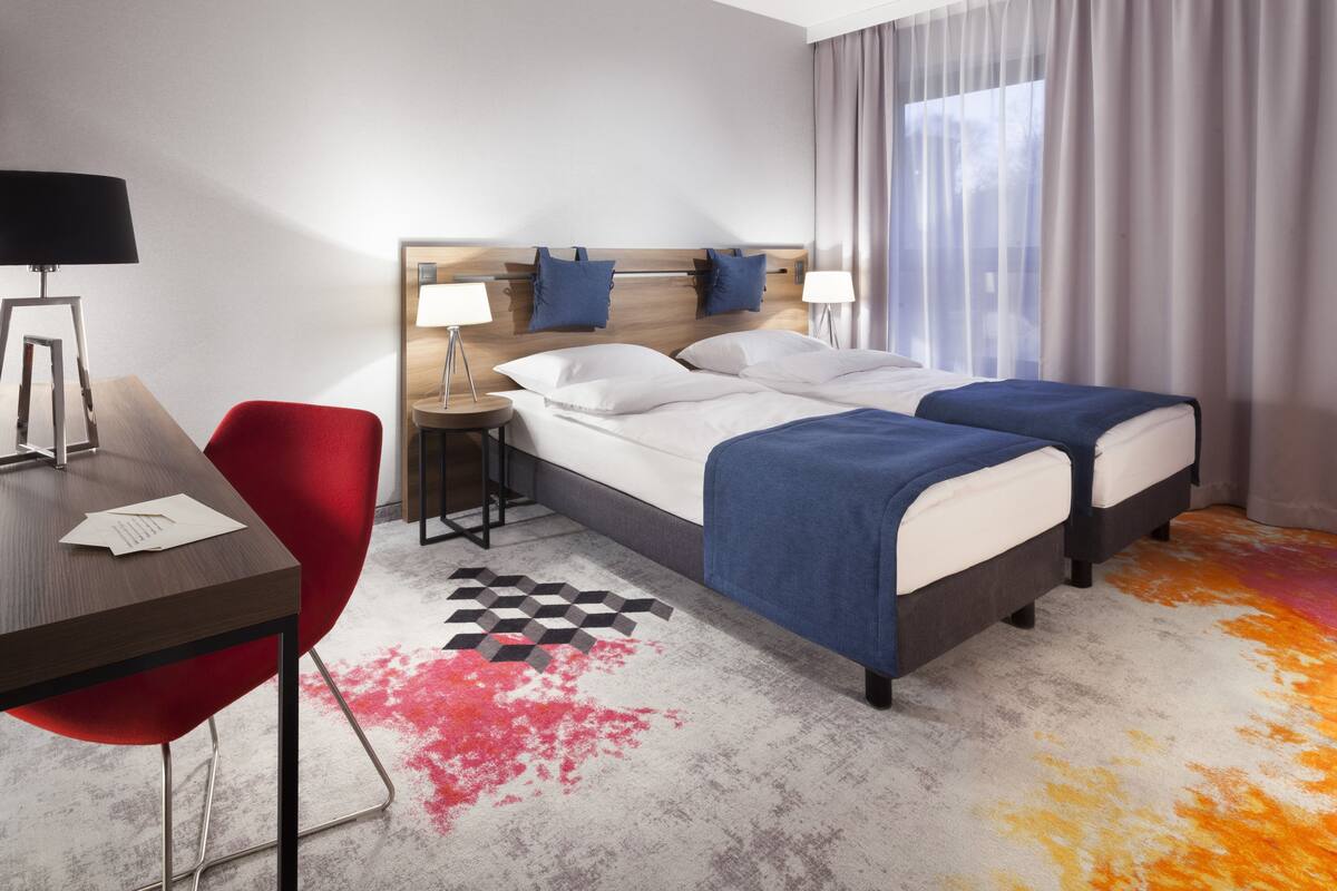 A hotel room with a bed, two bedside tables, a desk, a chair, and a large window with curtains.