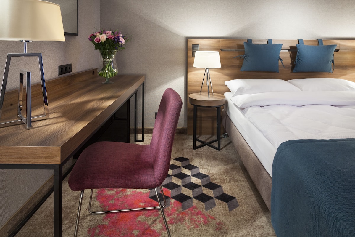 A hotel room with a wooden desk, a purple chair, a small round table with a lamp, a bed with white linens, and a vase with flowers on the desk.