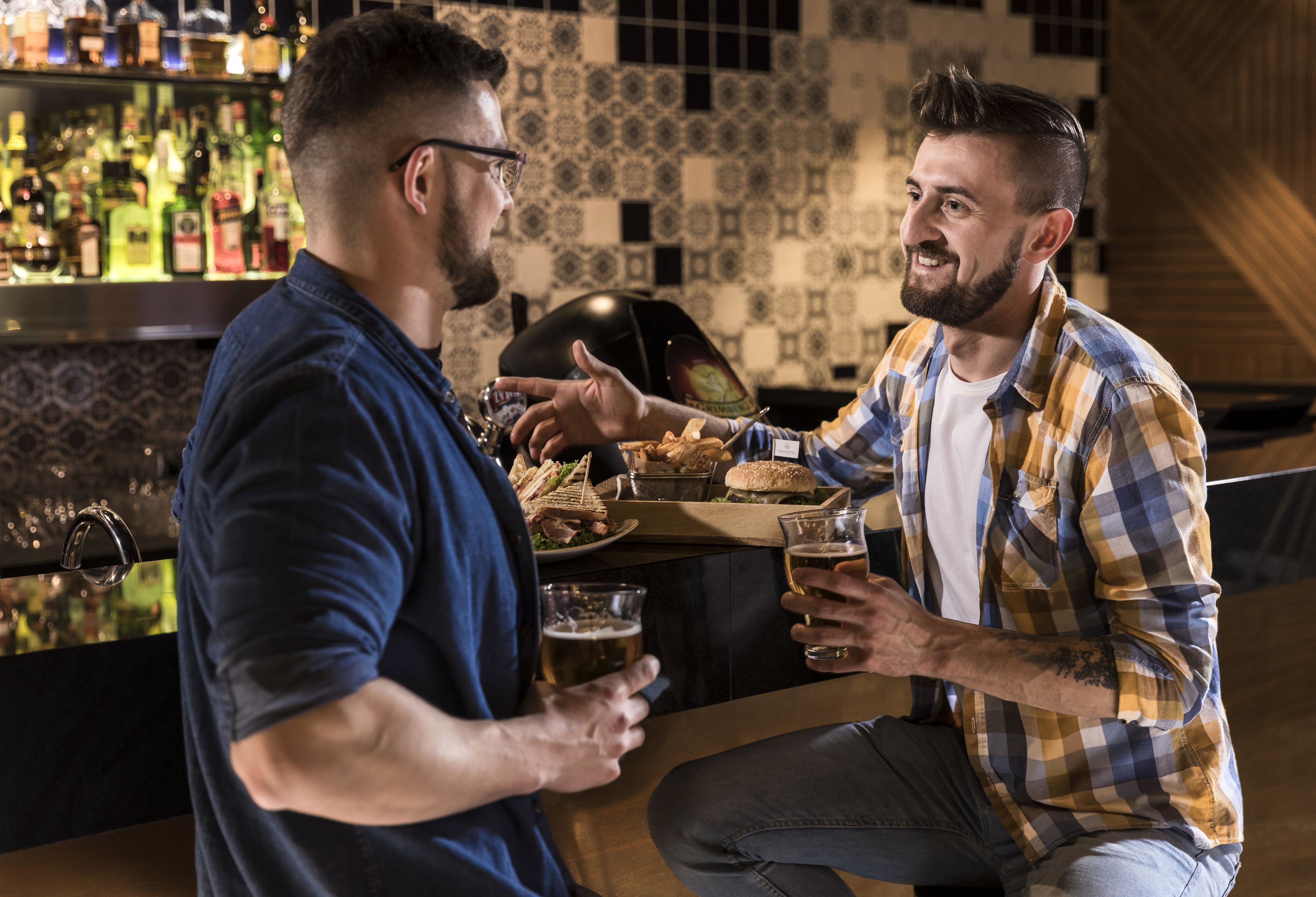 Two men sitting at a bar with a variety of drinks and snacks.
