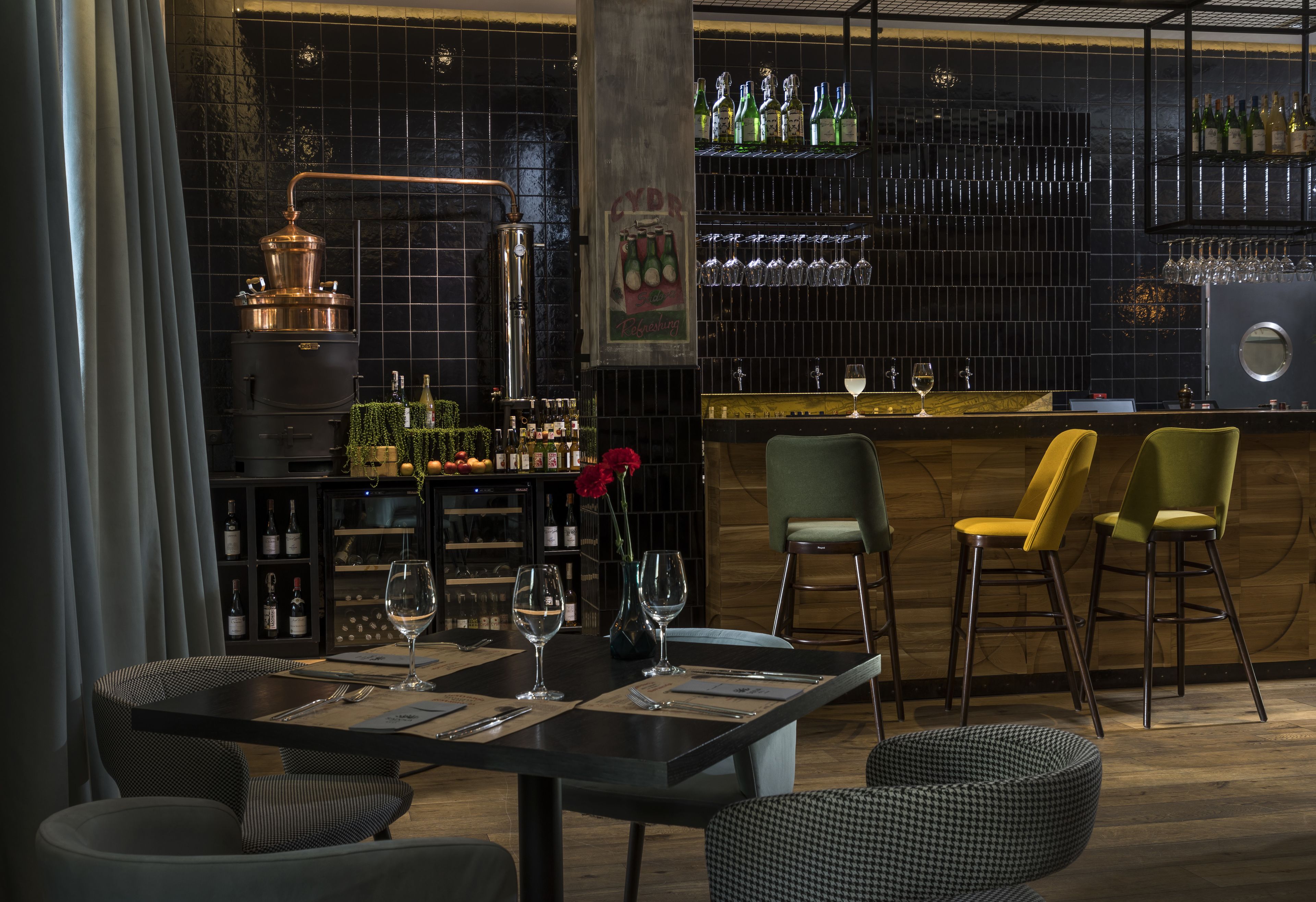 Un espace bar avec un comptoir, des étagères remplies de bouteilles et de verres, ainsi qu’un appareil de distillation en cuivre.