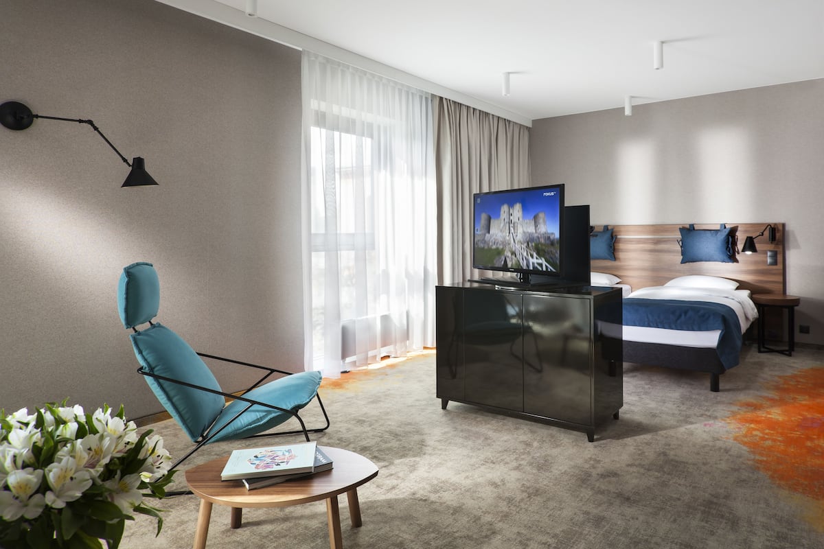 A modern hotel room with a bed, a TV, a desk, a chair, and a large window with curtains.