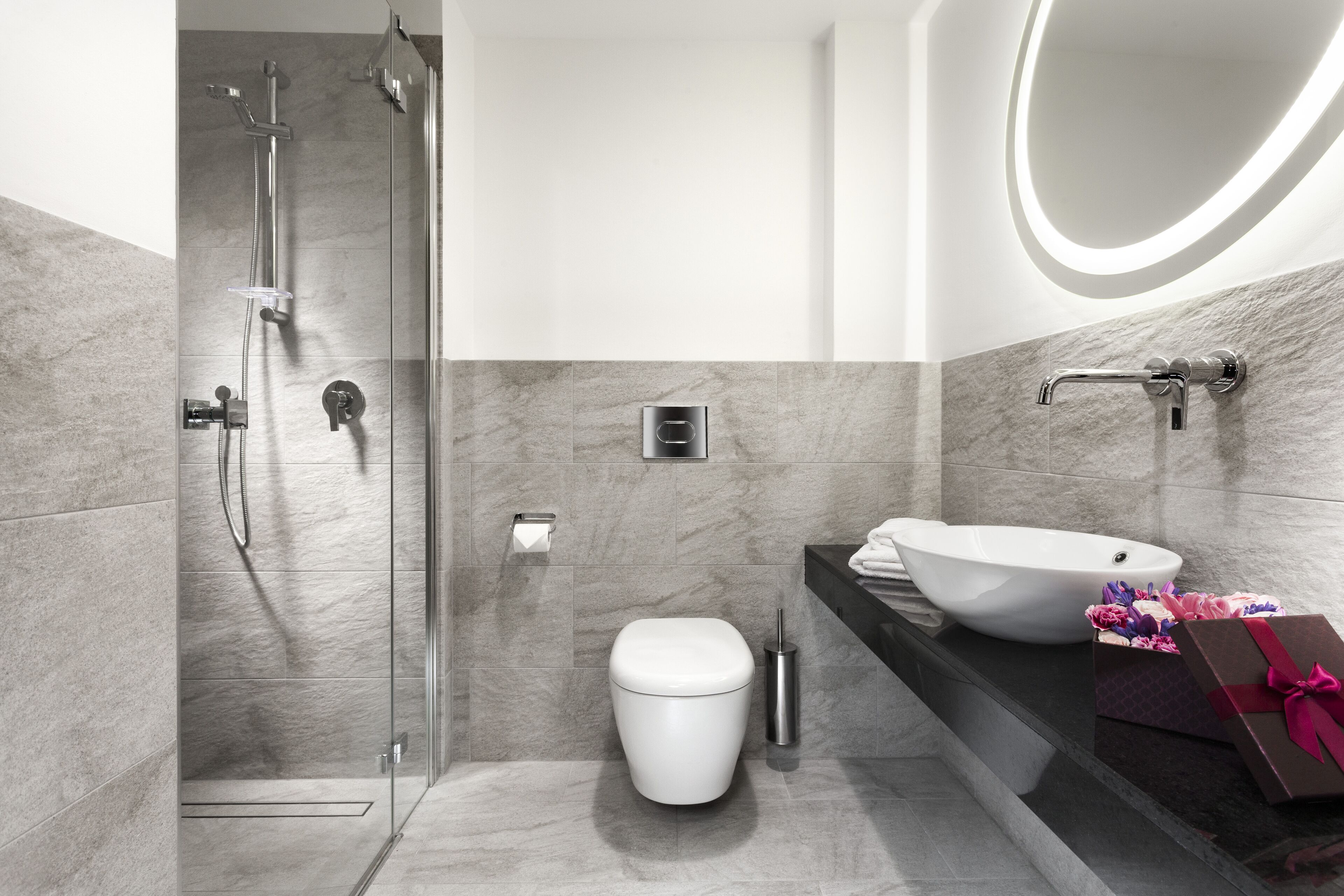 A modern bathroom with a glass-enclosed shower, a white toilet, a marble-tiled wall, a white sink, and a gift box on the counter.