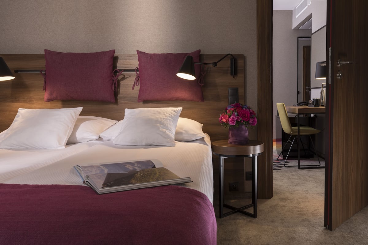 A hotel room with a bed, two pillows, a bedside table with a lamp, a small table with a vase of flowers, and a chair.