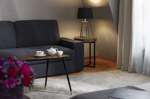 A modern living room with a dark sofa, a small round table with a teapot and cups, a lamp on a side table, and a potted plant.