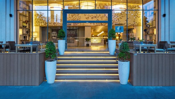 A modern hotel entrance with a glass facade, outdoor seating area, and a staircase leading to the lobby.