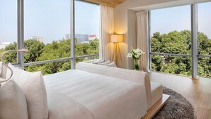 A modern hotel room with large windows, a bed, a bedside table with a lamp, and a view of the cityscape and greenery.