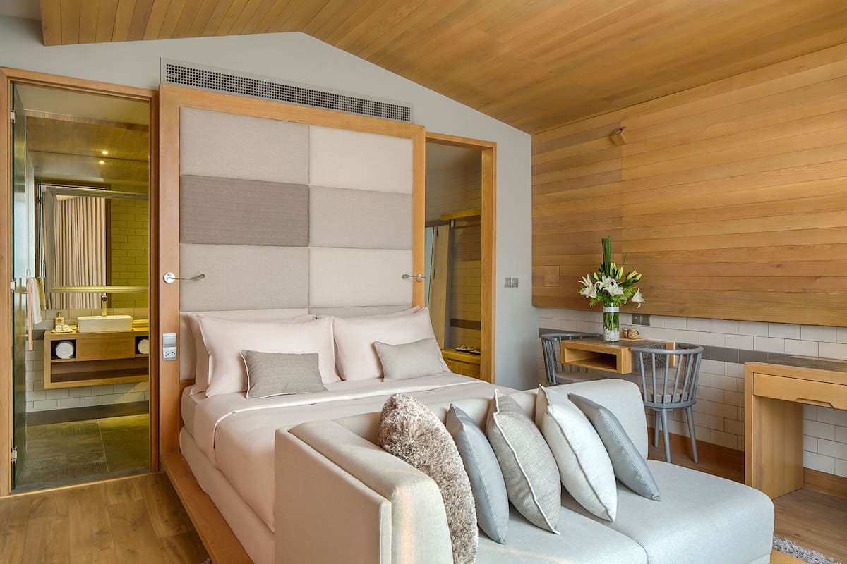 A modern bedroom with a large bed, a sofa, a small table, and a bathroom visible through a glass door.