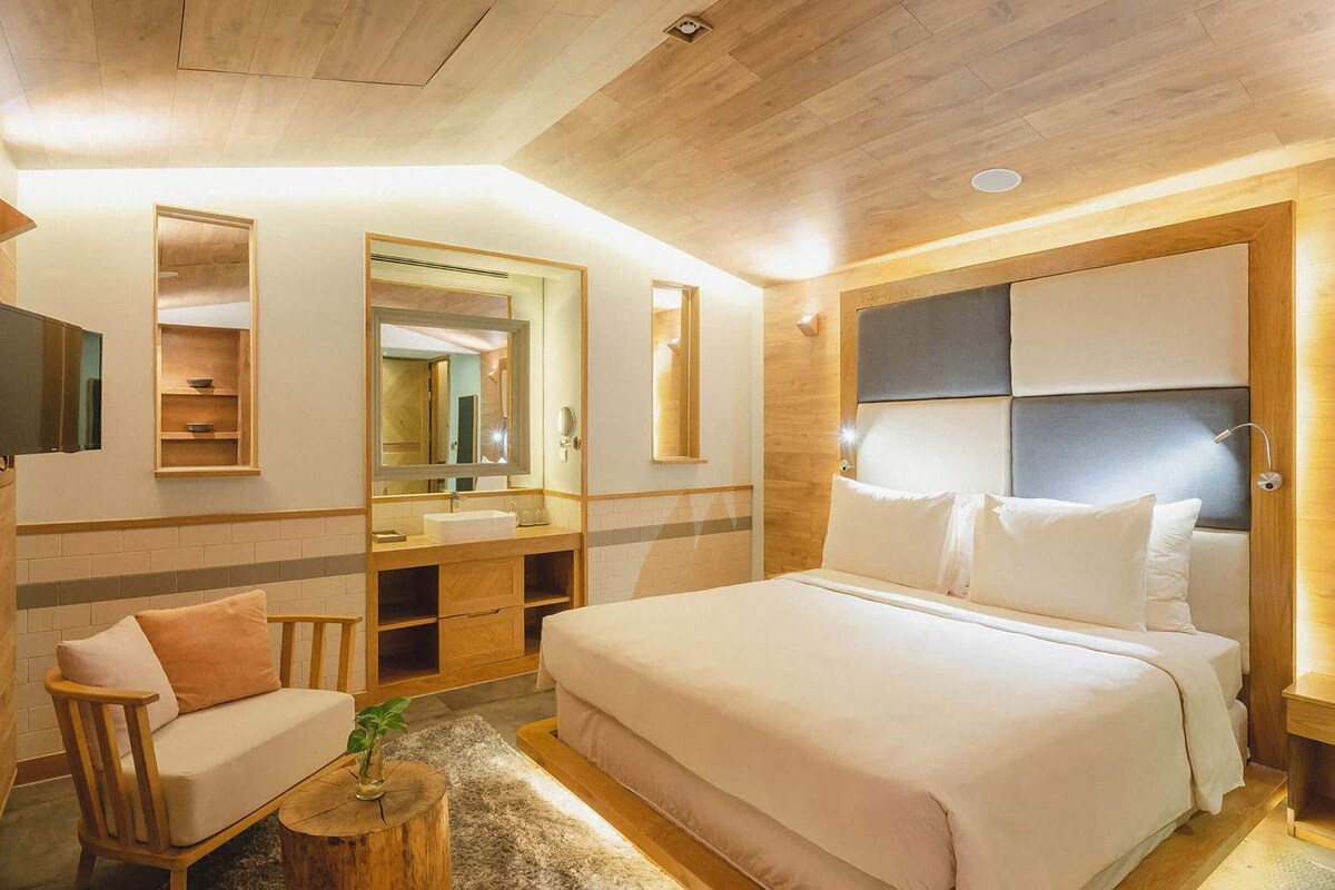 A hotel room with a bed, a wooden vanity, a mirror, a small seating area, and a nightstand.