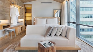 A modern bedroom with a large bed, a wooden headboard, a small wooden stool, and a view of a building through a large window.