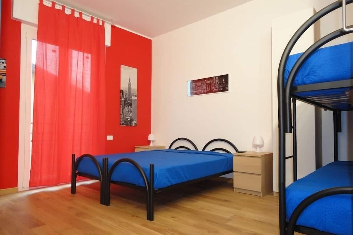 Une chambre avec deux lits simples, un lit superposé, un plancher en bois, un rideau rouge et un tableau représentant un paysage urbain accroché au mur.