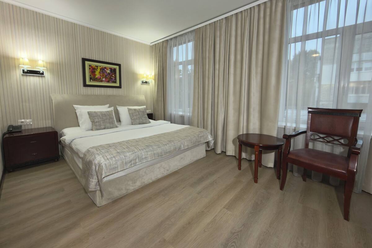 A hotel room with a large bed, a wooden chair, a small table, a nightstand, and a painting on the wall.