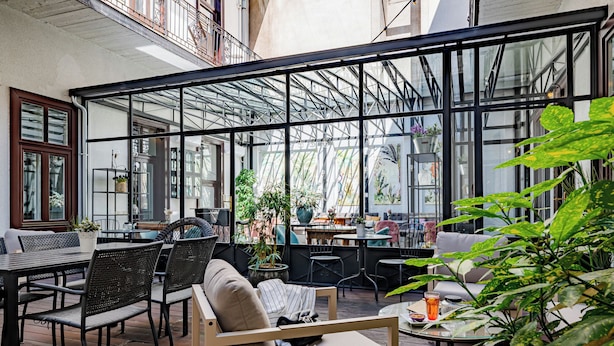 Un café en plein air avec des tables et des chaises, entouré de murs de verre et agrémenté de plantes en pot.