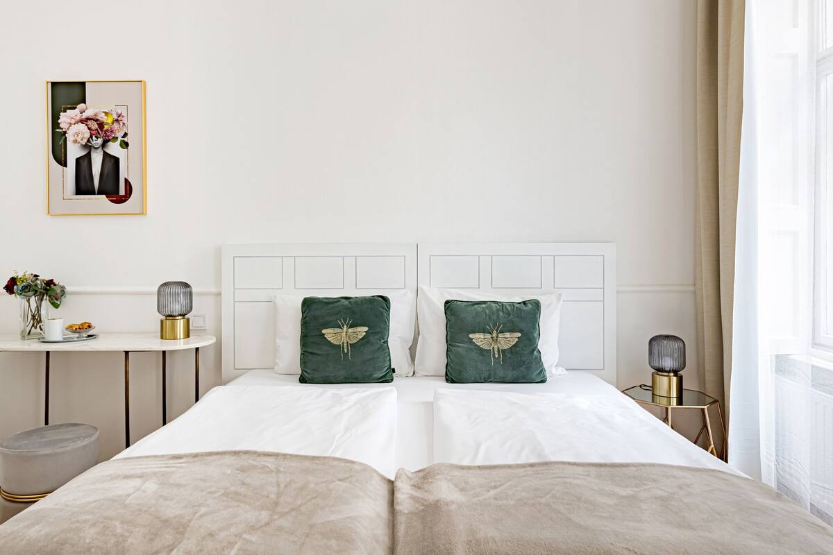 Une chambre à coucher bien rangée, avec un lit, deux oreillers ornés d’un motif en forme d’ailes, une table de chevet avec une lampe, une petite table ronde sur laquelle est posé un vase de fleurs, et un tableau encadré accroché au mur.