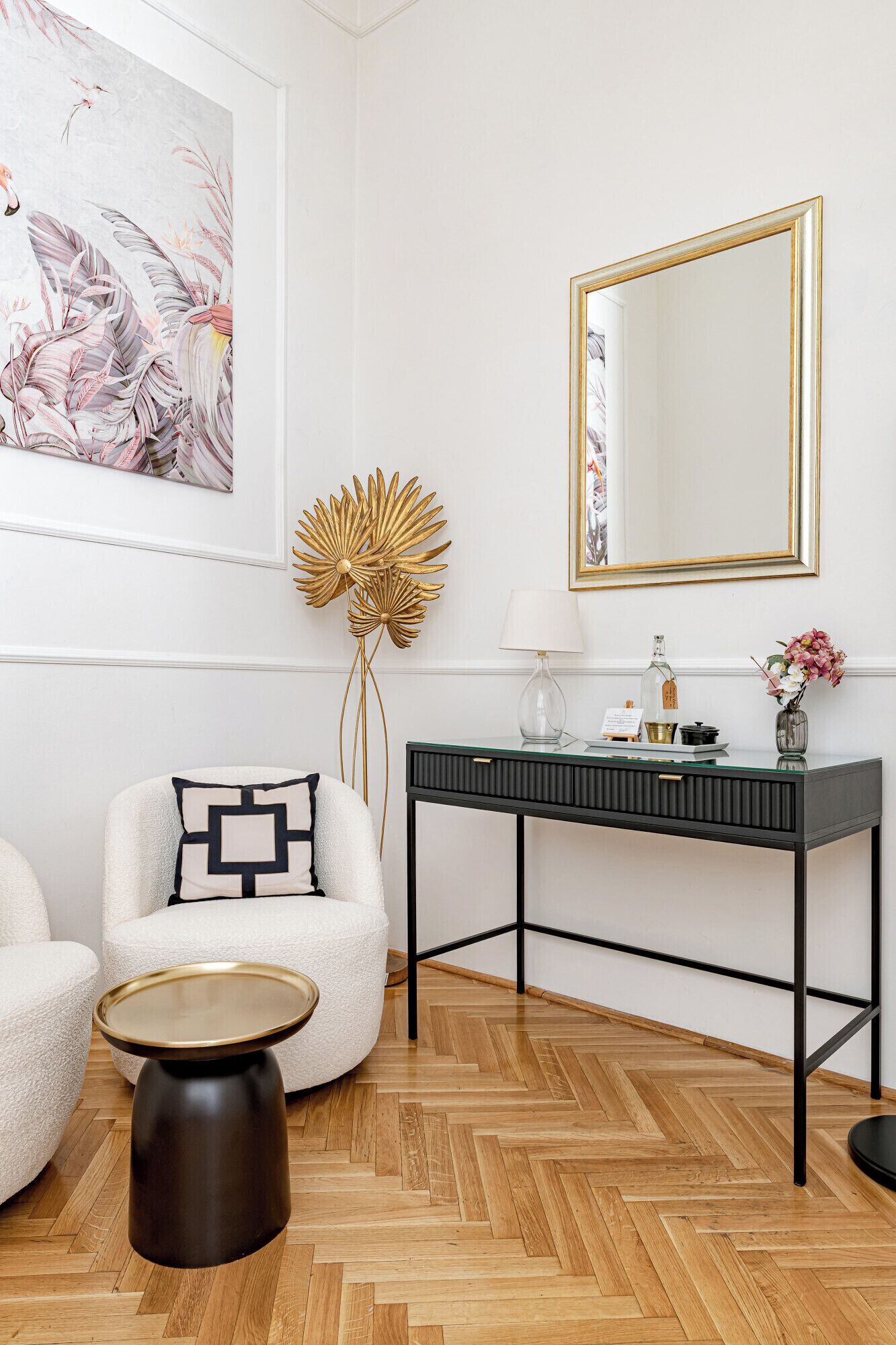 Una sala de estar moderna con piso de madera en patrón espiga, una mesa de centro redonda, un sillón blanco con un cojín de diseño geométrico, una consola negra con un espejo, y un cuadro abstracto grande en la pared.