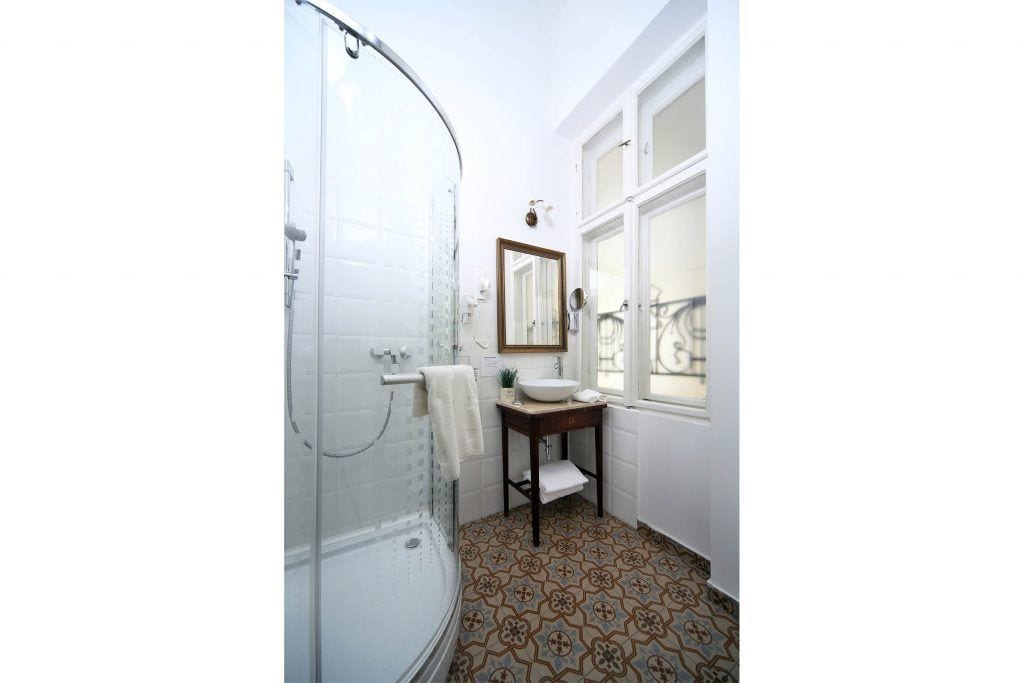 A bathroom with a glass shower enclosure, a sink with a mirror above it, patterned floor tiles, and a window providing natural light.