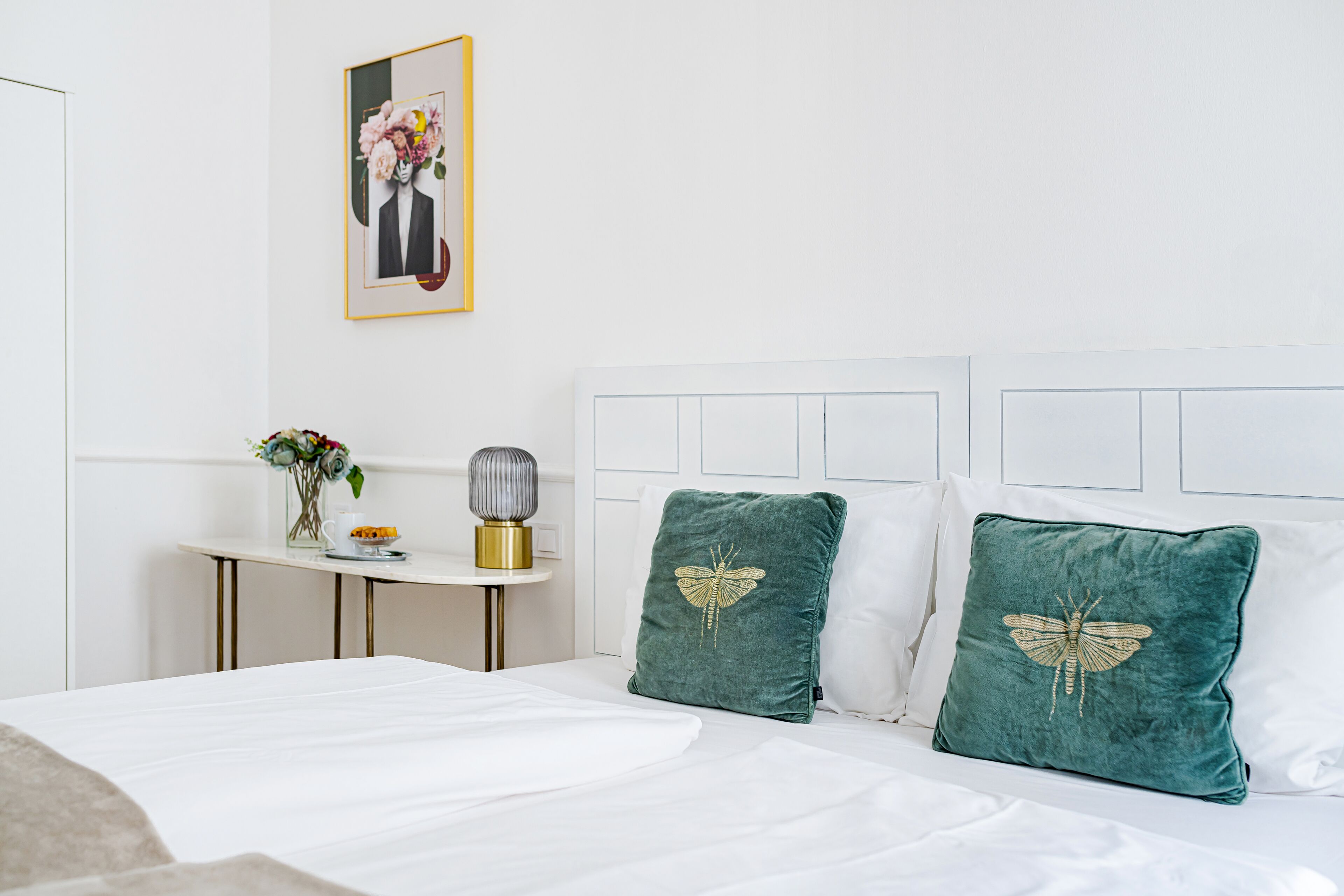 Una cama bien tendida con sábanas blancas, una mesita con un florero y un cuadro en la pared.