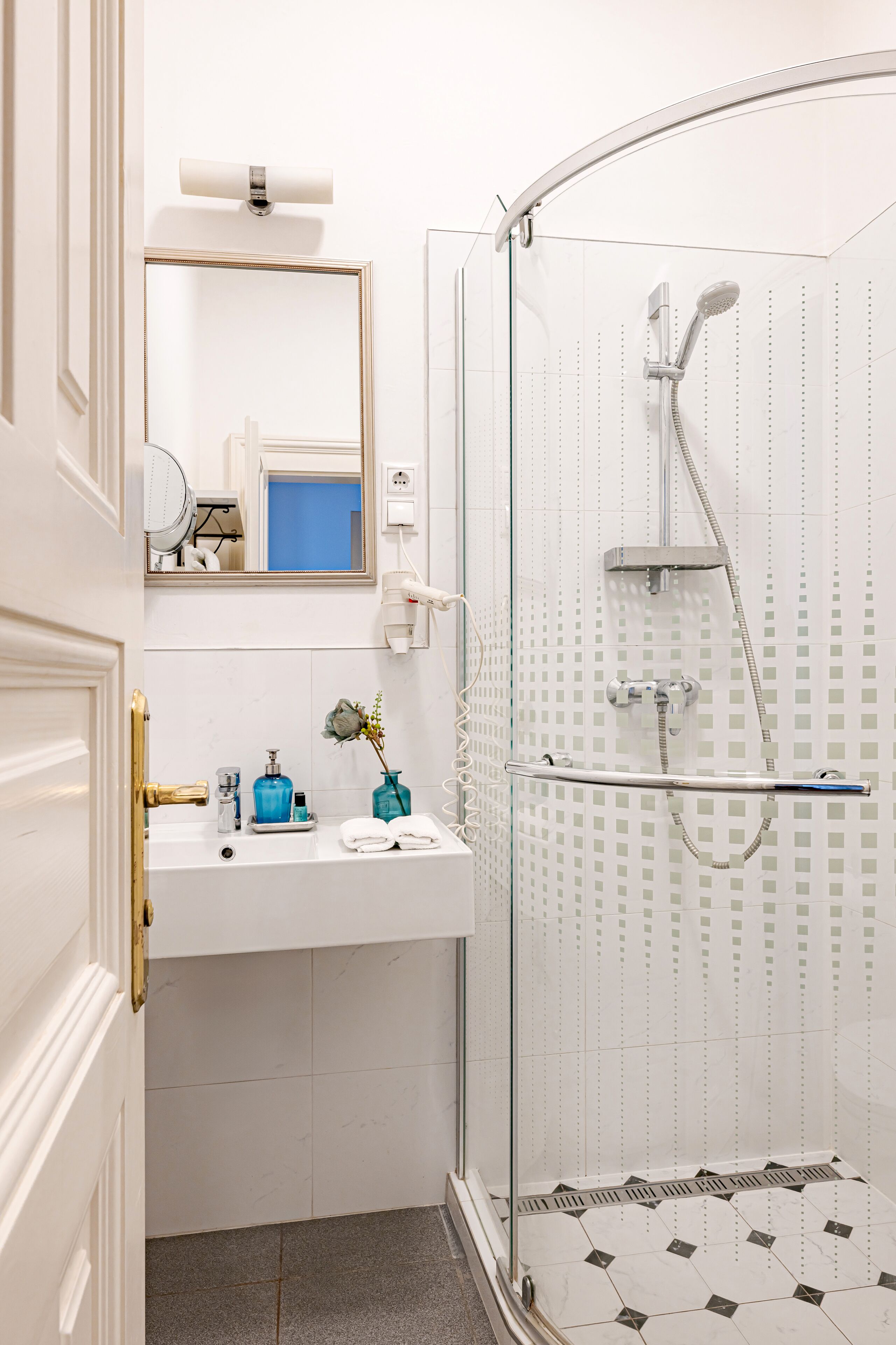 Une salle de bain avec une douche à parois de verre, un lavabo blanc avec un miroir au-dessus, et un petit vase avec des fleurs.