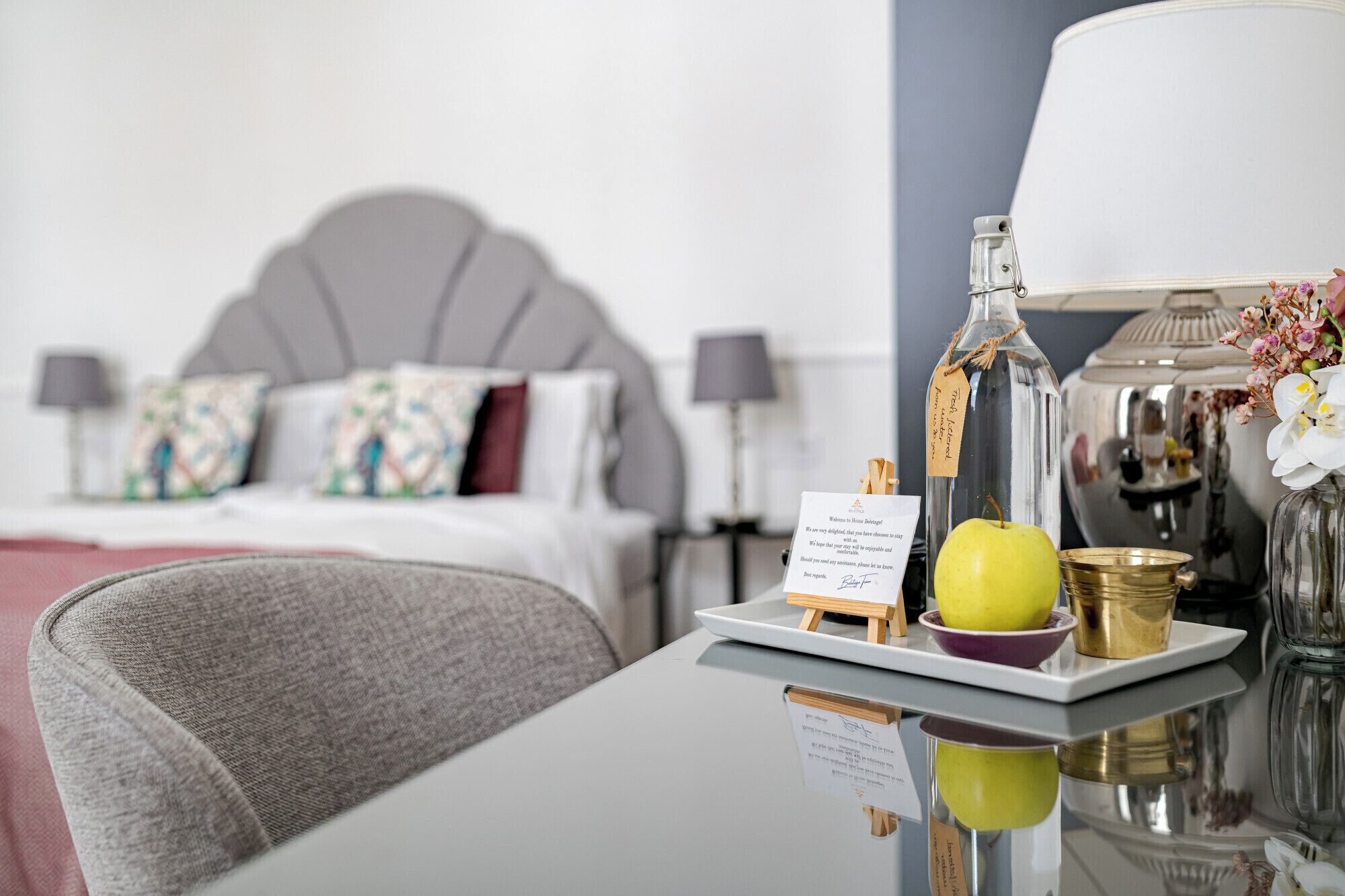 A hotel room with a neatly made bed, a bedside table with a lamp, a tray with a bottle, an apple, and a bowl.