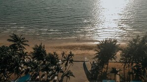 Une vue aérienne d’une plage bordée de palmiers, avec une étendue de sable et quelques bâtiments aux toits plats.