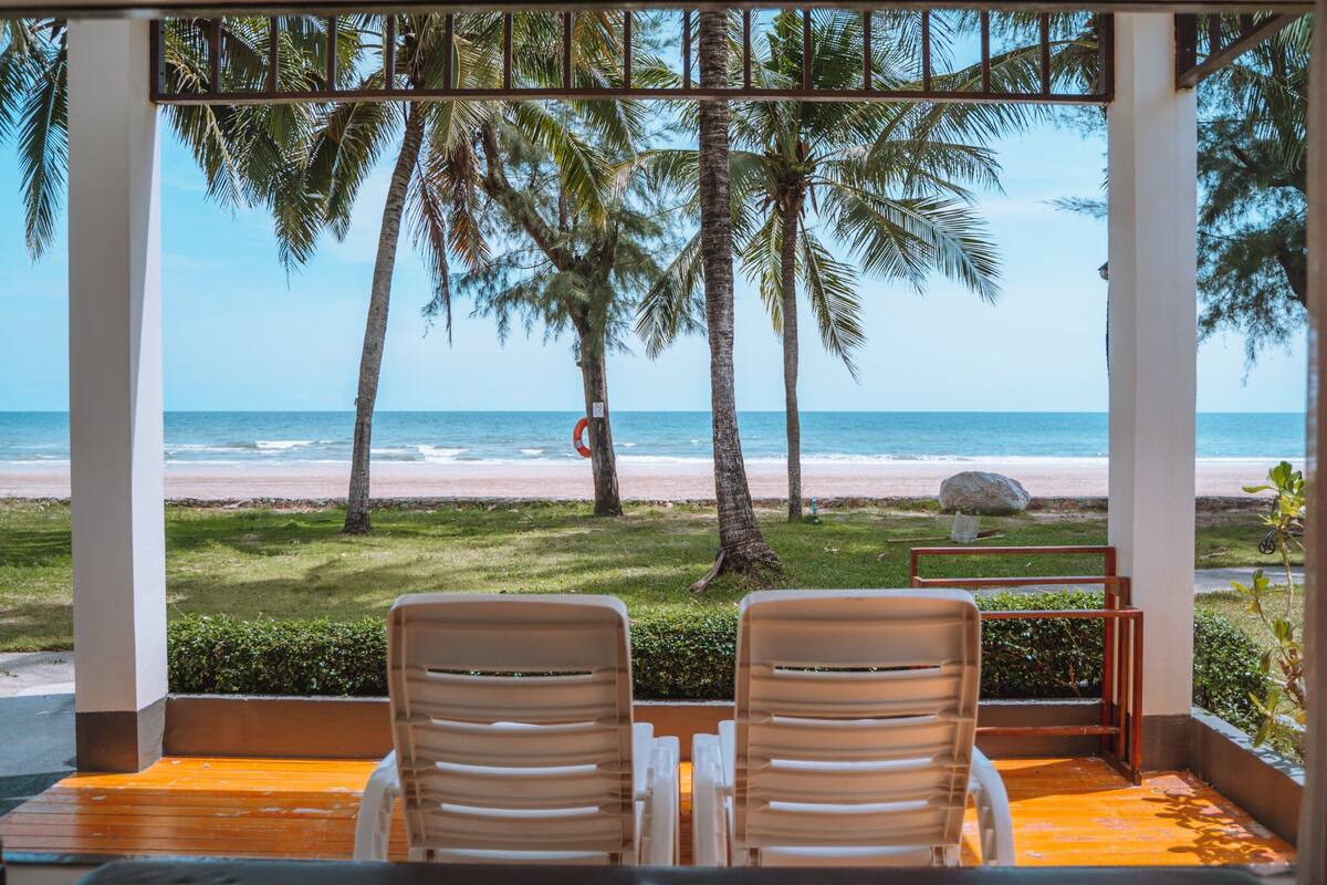 A covered outdoor seating area with two chairs facing a beach view, palm trees, and a lifeguard stand.