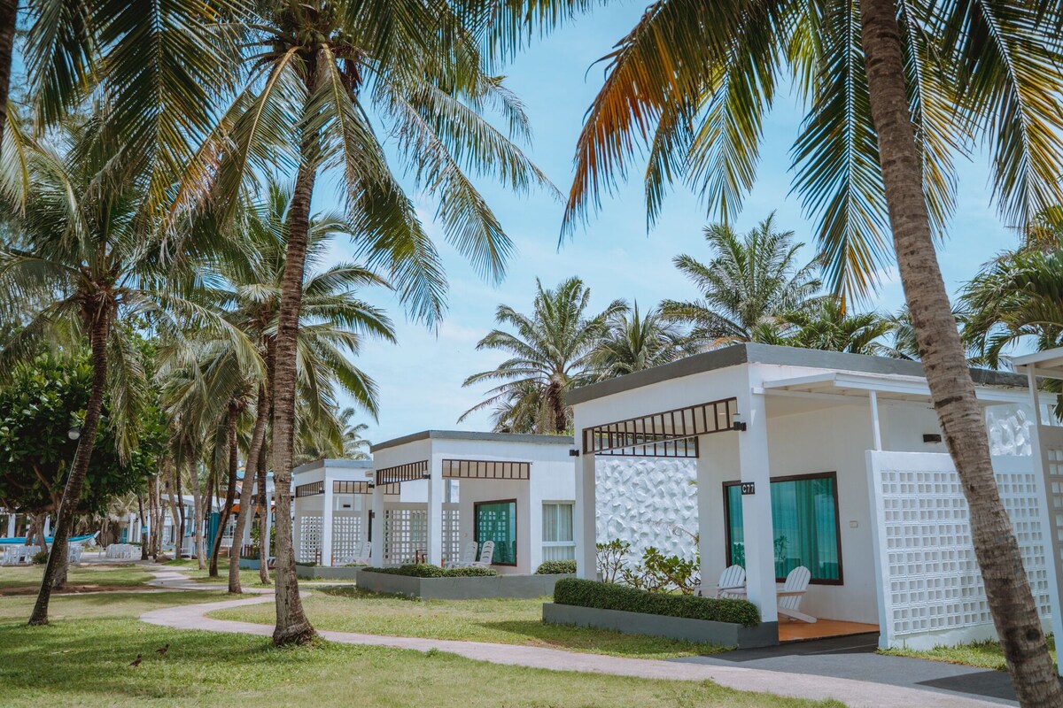 Une rangée de villas modernes aux façades blanches et aux portes vertes, entourées de palmiers et surplombées par un ciel d’un bleu limpide.