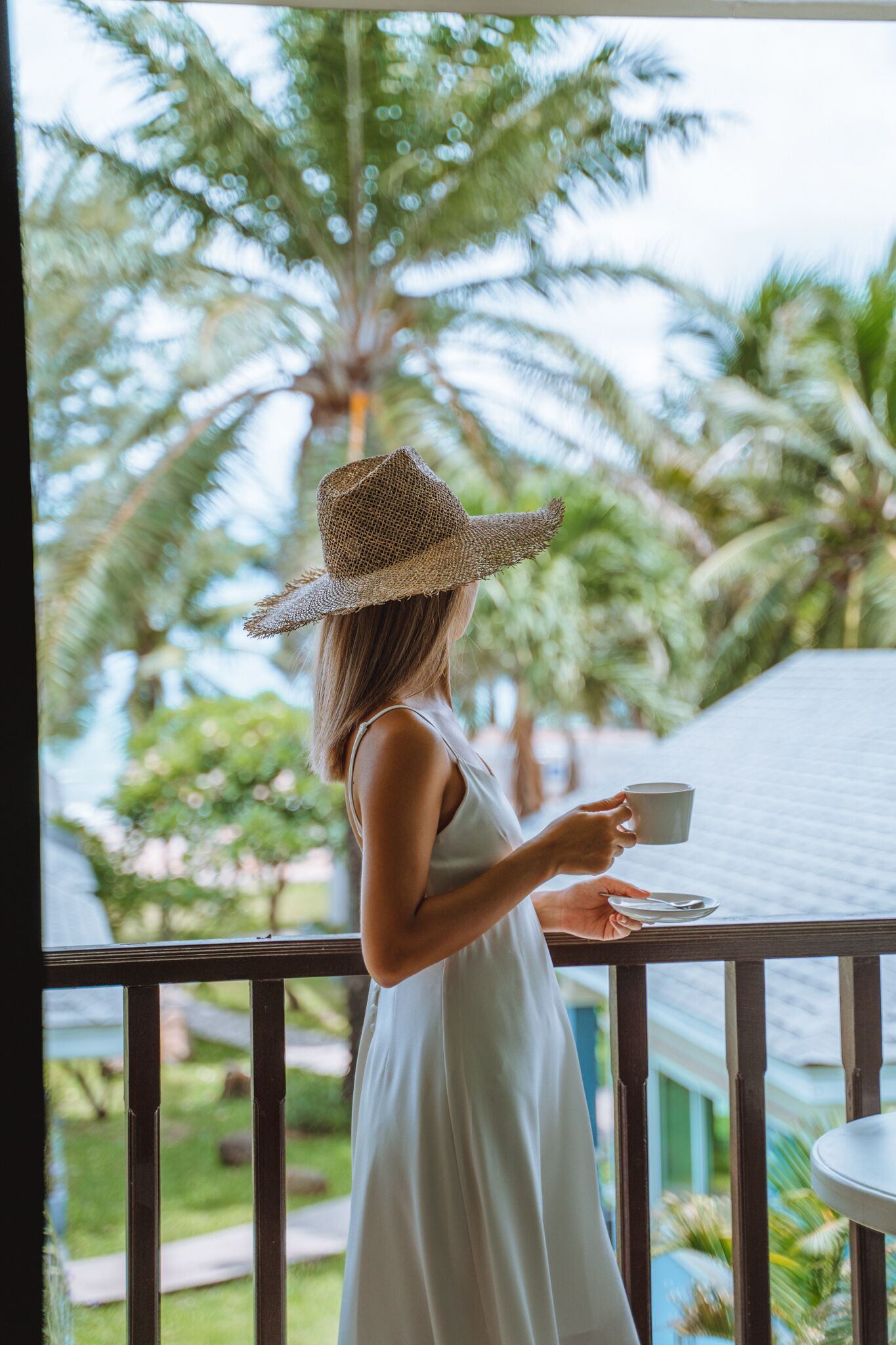 A person in a white dress standing on a balcony with a view of palm trees and a house.