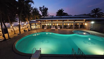 Una piscina con sillones de descanso y un bar, rodeada de palmeras y un edificio con amplios ventanales.