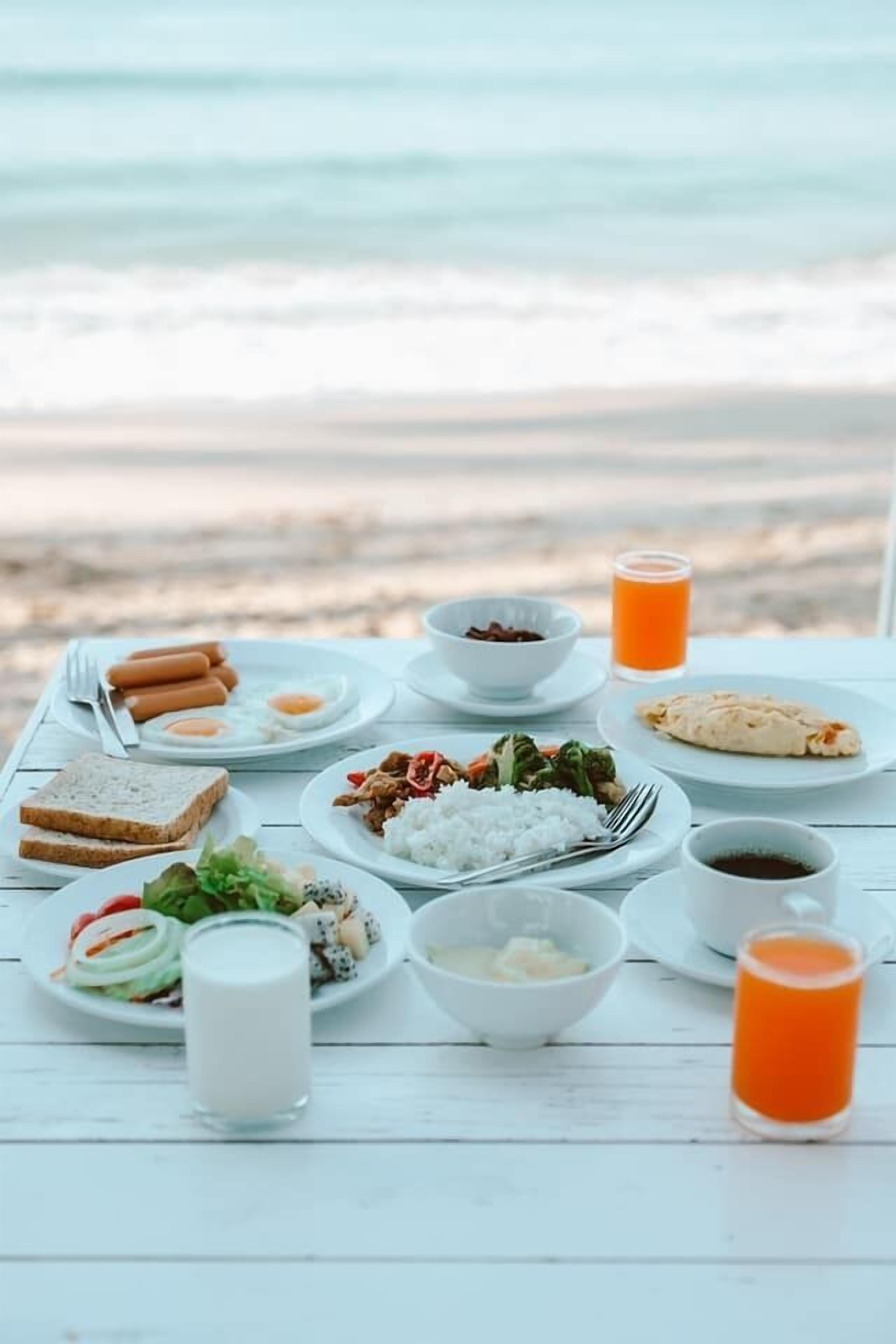 A table set with breakfast items including eggs, toast, rice, and vegetables, with a view of the beach in the background.