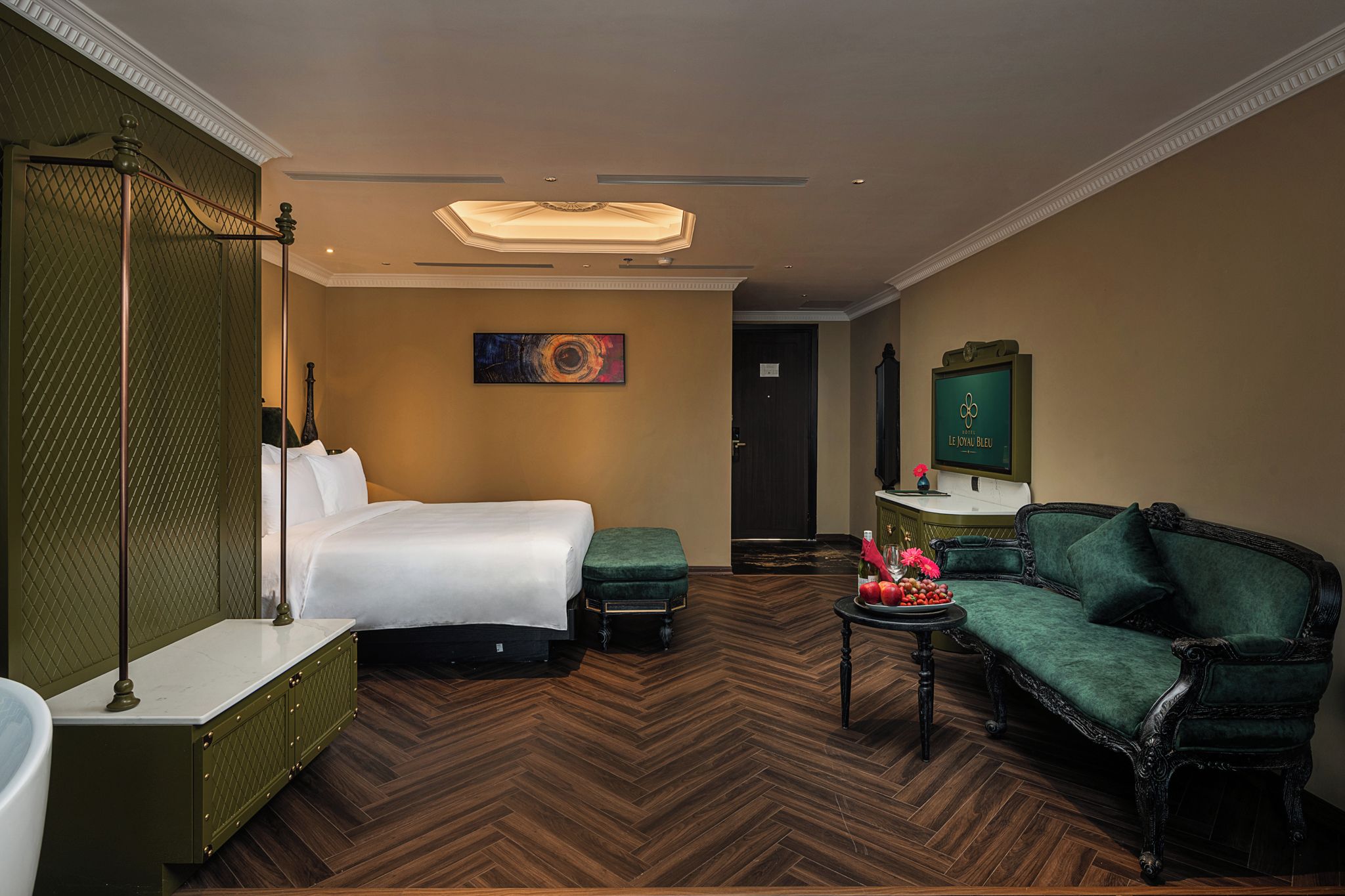 Une chambre d’hôtel avec un lit, un canapé vert, une petite table avec des fruits et un tableau au mur.