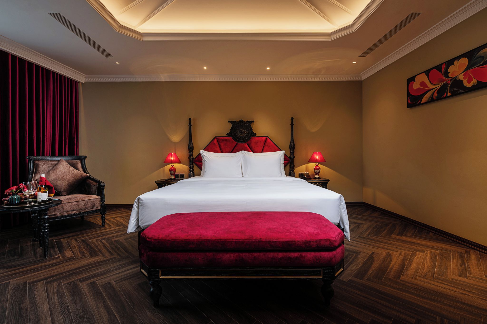 Una habitación de hotel con una cama grande, un banco tapizado de rojo, una mesita de noche de madera con lámparas y una cortina roja.
