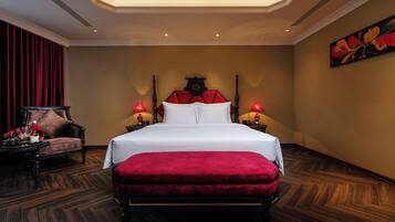 Una habitación de hotel con una cama grande, un banco tapizado de rojo, una mesita de noche de madera con lámparas y una cortina roja.