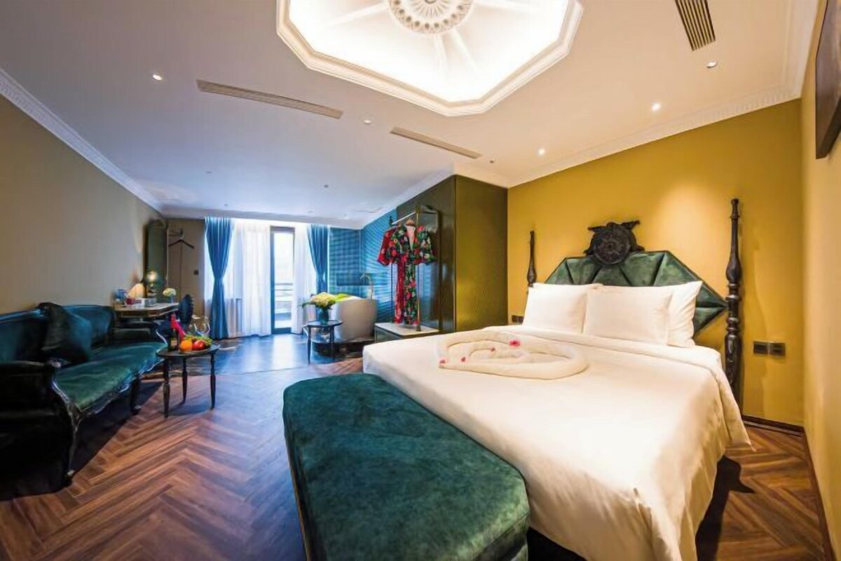 A hotel room with a large bed, a sitting area with a sofa and armchair, a desk with a chair, and a view of the outside.