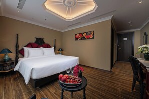 A hotel room with a large bed, a side table with fruit and glasses, a painting on the wall, and a wooden floor. - Hôtel Le Joyau Bleu Hanoï (Hanoi)