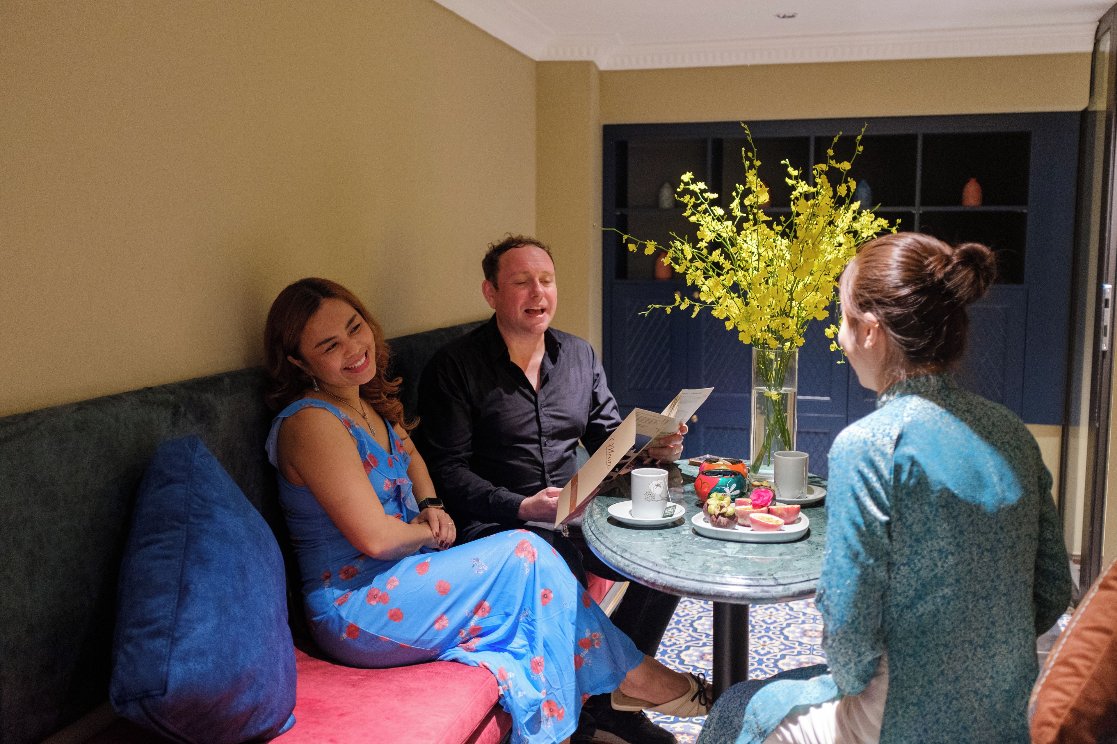 A group of people seated on a couch and a table with a vase of flowers and a plate of food.