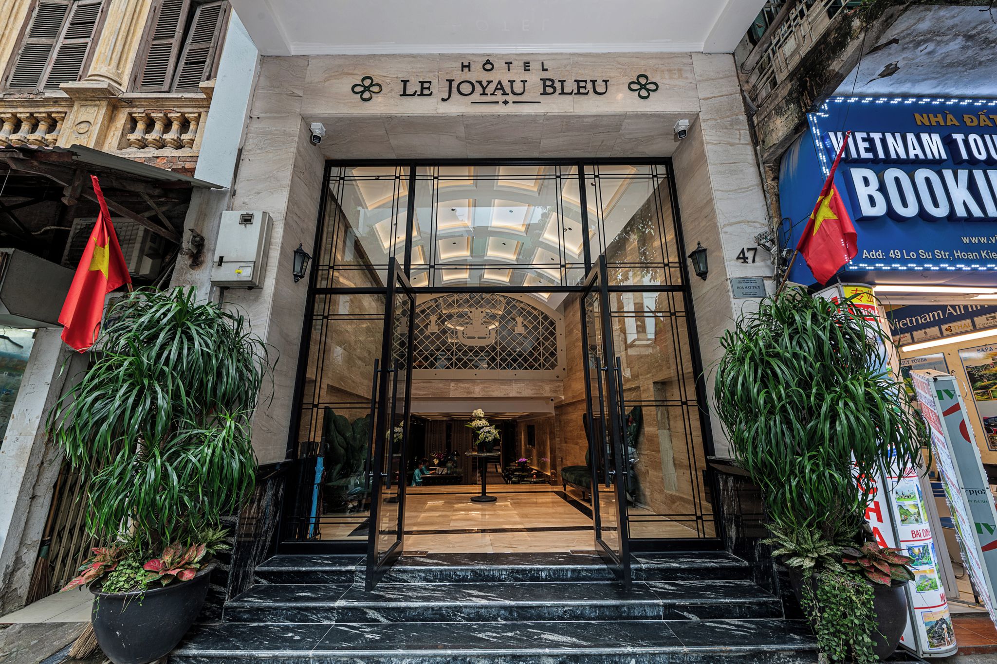 The entrance to Hotel Le Joyau Bleu with a glass facade, a central staircase, and a flag on the left.