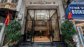 The entrance to Hotel Le Joyau Bleu with a glass facade, a central staircase, and a flag on the left.