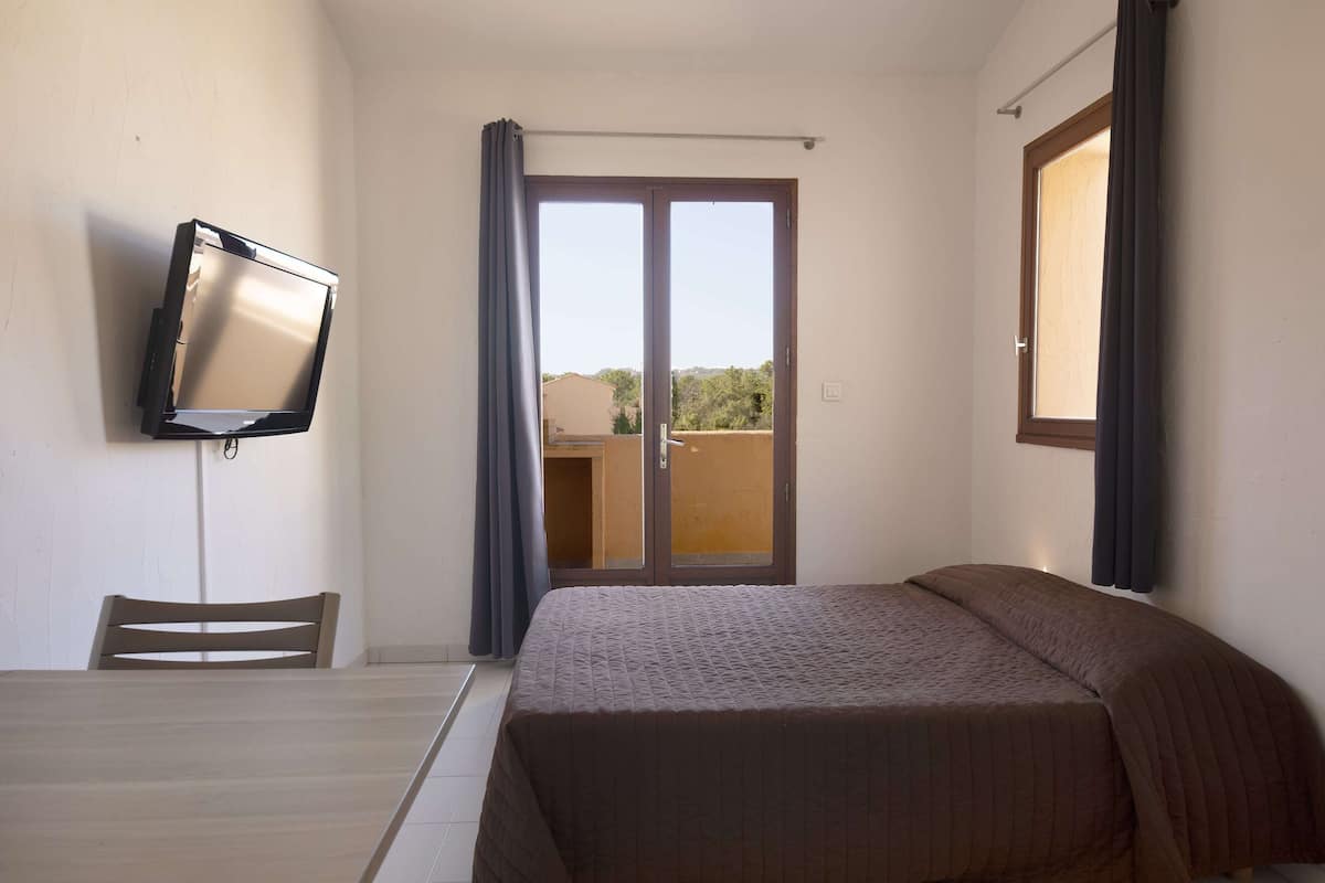 A hotel room with a bed, a TV mounted on the wall, a dining table with chairs, and a sliding glass door leading to an outdoor area.