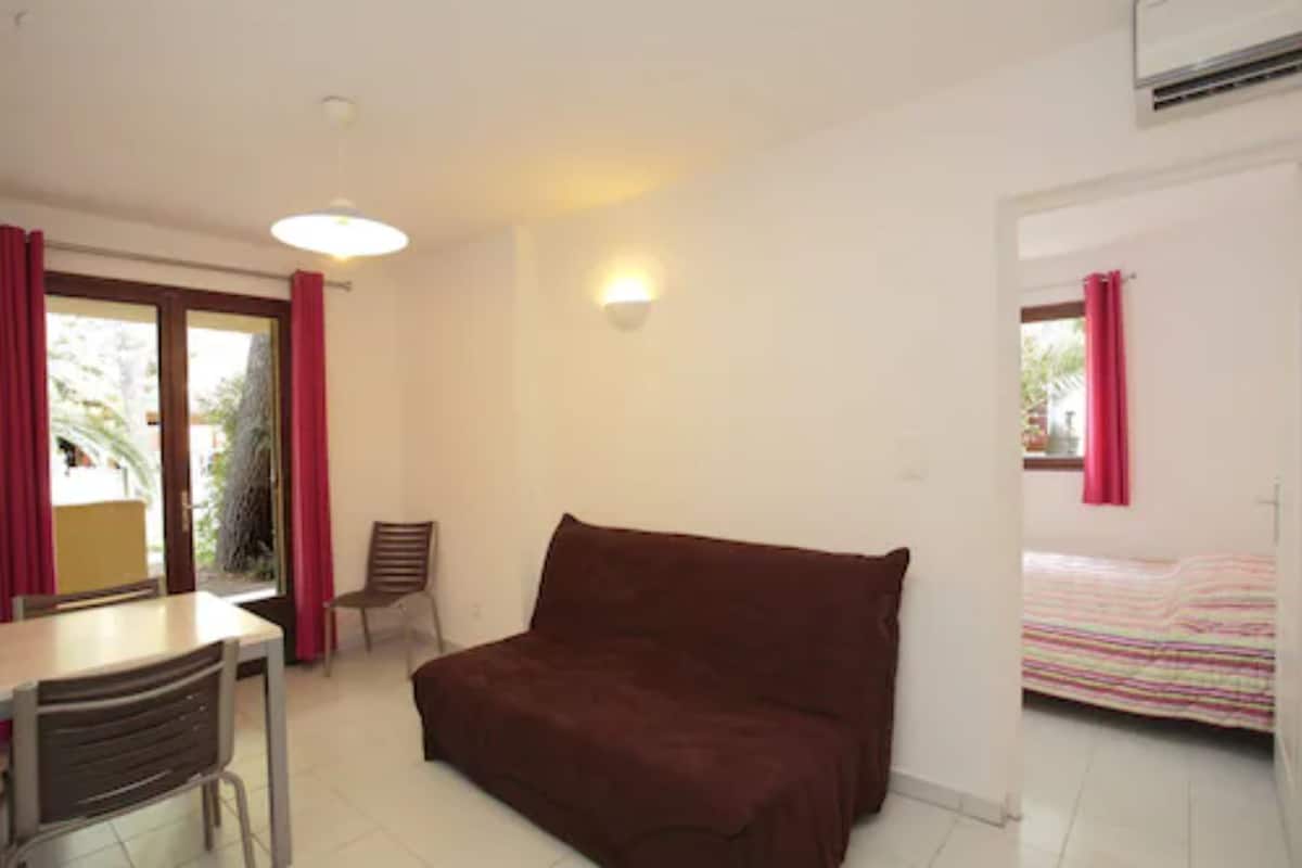 A room with a brown sofa, a dining table with chairs, a small table, a wall-mounted light, and a bed with a striped blanket.